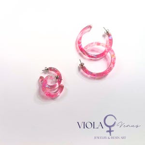 May include: Two pairs of translucent pink resin hoop earrings featuring embedded iridescent glitter flakes. Each hoop has a silver-toned metal post backing. The text VIOLA Venus Jewelry & Resin Art with a female symbol is printed in the bottom right corner.