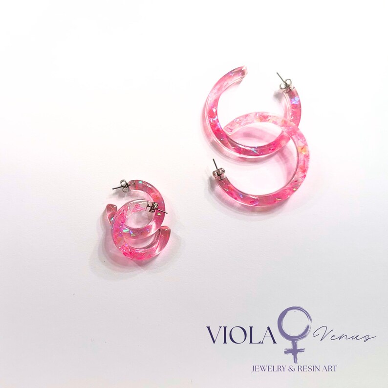 May include: Two pairs of translucent pink resin hoop earrings featuring embedded iridescent glitter flakes. Each hoop has a silver-toned metal post backing. The text VIOLA Venus Jewelry & Resin Art with a female symbol is printed in the bottom right corner.