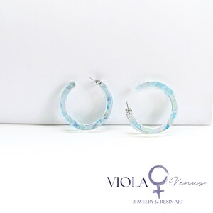 May include: A pair of translucent hoop earrings featuring embedded iridescent blue and purple flakes. The earrings have a circular shape with a silver-toned metal post. The text VIOLA Venus JEWELRY & RESIN ART is printed in the bottom right corner.