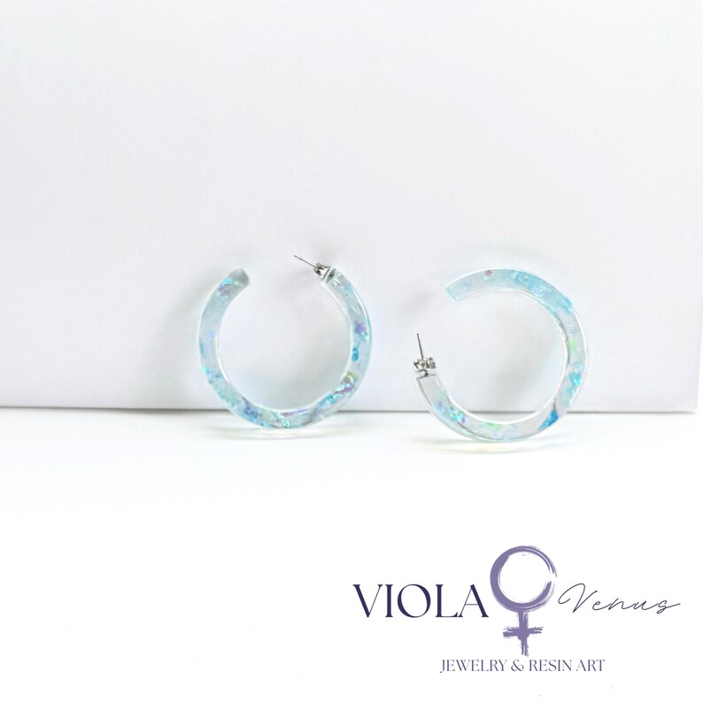 May include: A pair of translucent hoop earrings featuring embedded iridescent blue and purple flakes. The earrings have a circular shape with a silver-toned metal post. The text VIOLA Venus JEWELRY & RESIN ART is printed in the bottom right corner.
