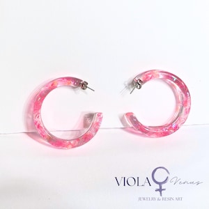 May include: A pair of clear resin hoop earrings featuring embedded pink and iridescent flakes. The earrings have silver-toned metal posts and backs. The text VIOLA Venus JEWELRY & RESIN ART with a female symbol is printed in the bottom right corner.