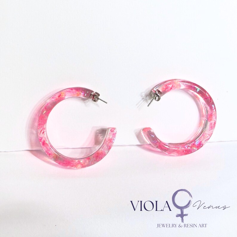May include: A pair of clear resin hoop earrings featuring embedded pink and iridescent flakes. The earrings have silver-toned metal posts and backs. The text VIOLA Venus JEWELRY & RESIN ART with a female symbol is printed in the bottom right corner.
