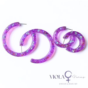 May include: Two pairs of purple resin hoop earrings featuring embedded iridescent flakes that catch the light. The earrings have a translucent finish and include metal posts. The text VIOLA Venus JEWELRY & RESIN ART is visible.