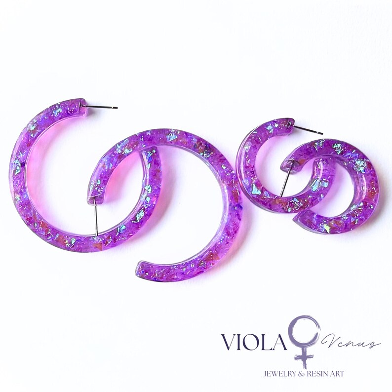 May include: Two pairs of purple resin hoop earrings featuring embedded iridescent flakes that catch the light. The earrings have a translucent finish and include metal posts. The text VIOLA Venus JEWELRY & RESIN ART is visible.