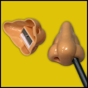 May include: Novelty pencil sharpener shaped like a human nose, in a light tan color. The sharpener is open, revealing the blade and pencil shavings. A pencil is inserted into the other nose-shaped piece.