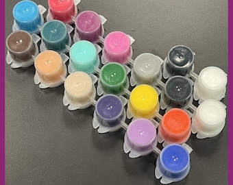 May include: An assortment of colorful tattoo ink cups in various shades, including blue, red, green, and yellow. The ink cups are arranged in a grid pattern on a dark surface, with a purple border.