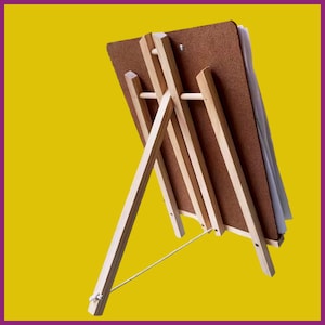 May include: A wooden artist's easel with a brown backing board and a stack of white paper. The easel is made of light-colored wood and has a simple, functional design. The background is a solid yellow.
