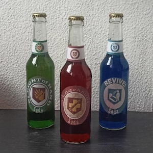 Könnte beinhalten: Drei Glas-Limonadenflaschen mit goldenen Verschlüssen. Die Flaschen enthalten grüne, rote und blaue Flüssigkeiten. Jede Flasche hat ein Etikett mit einem kreisförmigen Design und dem Text: "LIGHT OF HAND Soda", "Juggernaut Soda" und "REVIVE Soda".