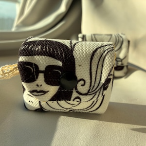May include: A small, rectangular, off-white fabric pouch featuring a black graphic print of a face wearing large sunglasses. The pouch has a black circular snap button closure on the front flap and a small, light-colored lace loop on the side.
