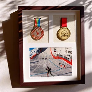 Könnte beinhalten: Eine gerahmte Präsentation mit zwei Medaillen und einem Foto einer Skipiste. Die Medaillen haben farbige Bänder. Das Foto zeigt Skifahrer auf einer schneebedeckten Piste mit einem Skilift und dem Wort "Garmisch".