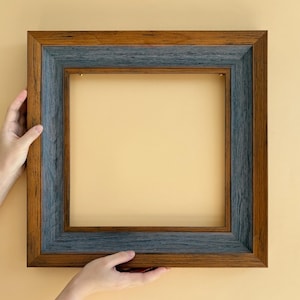 Pode incluir: Uma moldura quadrada de madeira com um design de duas cores. A moldura externa é marrom quente, enquanto a borda interna é cinza-azulada suave. A moldura é segurada por duas mãos contra um fundo amarelo claro.