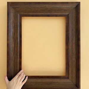Könnte beinhalten: Ein dunkelbrauner Holzbilderrahmen mit rechteckiger Öffnung. Der Rahmen hat eine strukturierte Oberfläche und wird von einer Hand gehalten. Der Hintergrund ist einfarbig hellgelb.