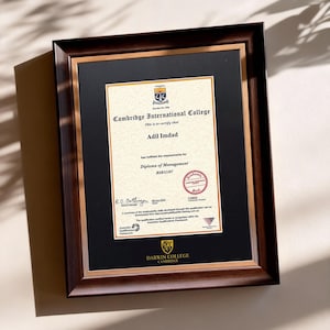 May include: A framed certificate from Cambridge International College. The document, with the name Adil Imdad, is presented in a dark brown wooden frame with a gold inner border. The text includes the college name, the title "Diploma of Management", and a seal.