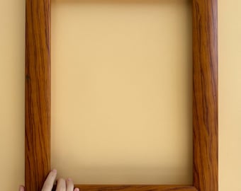 May include: A rectangular wooden picture frame with a warm brown finish. The frame has a visible wood grain pattern and is held by a hand against a light yellow background. The frame is empty, ready for a photo or artwork.