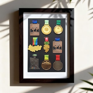 Könnte beinhalten: Ein gerahmtes Display verschiedener Medaillen, jede mit einem einzigartigen Design und Band. Die Medaillen sind in Gold-, Bronze- und Silbertönen gehalten und vor einem schwarzen Hintergrund in einem dunklen Holzrahmen angeordnet. Die Medaillen scheinen für sportliche Leistungen zu sein.