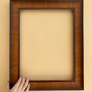 Könnte beinhalten: Ein rechteckiger Bilderrahmen aus Holz mit dunkelbraunem Finish. Der Rahmen hat ein schlichtes, klassisches Design und wird von einer Hand gehalten. Der Rahmen ist leer, bereit für ein Foto oder ein Kunstwerk.