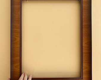 May include: A rectangular wooden picture frame with a dark brown finish. The frame has a simple, classic design and is held by a hand. The frame is empty, ready for a photo or artwork.