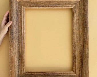May include: A rectangular wooden picture frame with a wide, textured border. The frame is held by two hands against a yellow background. The wood has a natural, rustic appearance with varying shades of brown.