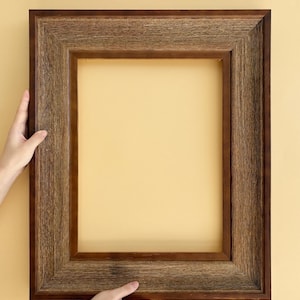 Op de afbeelding: Een rechthoekige, donkerbruine houten fotolijst met een getextureerde, rustieke uitstraling. Het frame wordt door twee handen tegen een lichtgele achtergrond gehouden. De binnenkant van het frame is leeg, klaar voor een foto of kunstwerk.