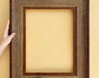 May include: A rectangular, dark brown wooden picture frame with a textured, rustic appearance. The frame is held by two hands against a light yellow background. The frame's interior is empty, ready for a photo or artwork.