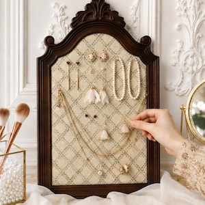 Könnte beinhalten: Ein dunkelbraunes, verziertes Schmuck-Display-Board mit gemustertem Stoffrücken. Das Board enthält verschiedene Ohrringe, Halsketten und ein Armband. Der Schmuck umfasst Perlen, Goldketten und weiße Blumenakzente. Eine Hand greift nach dem Schmuck.
