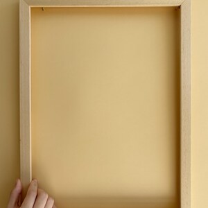 Könnte beinhalten: Ein rechteckiger, hellfarbiger Holzbilderrahmen. Der Rahmen ist leer, mit einem hellgelben Hintergrund. Eine Hand hält den Rahmen auf der linken Seite. Der Rahmen ist aus Naturholz.