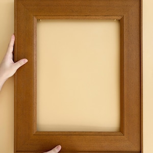 Könnte beinhalten: Ein rechteckiger Bilderrahmen aus Holz mit warmbraunem Finish. Der Rahmen wird von zwei Händen vor einem hellgelben Hintergrund gehalten. Das Design des Rahmens weist eine leicht erhöhte Innenkante auf.