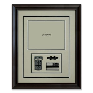 May include: A framed display with a dark brown wooden frame. The frame holds a photo opening and three military patches: a Ranger tab, a black and white American flag, and a military insignia. The background is a light beige.