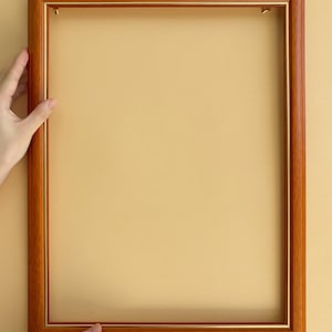 Puede incluir: Un marco de fotos rectangular de madera con un acabado marrón cálido y un fino borde interior dorado. El marco está sostenido por dos manos sobre un fondo amarillo claro, mostrando sus líneas limpias y su diseño clásico.