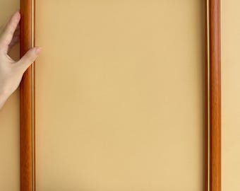 May include: A rectangular wooden picture frame with a warm brown finish and a thin gold inner border. The frame is held by two hands against a light yellow background, showcasing its clean lines and classic design.