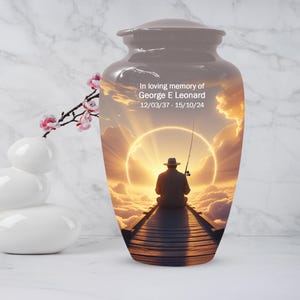 May include: A gray cremation urn with a serene scene of a person fishing on a pier at sunset. The urn has the text "In loving memory of George E Leonard 12/03/37 - 15/10/24". A white vase with pink flowers is on the left.