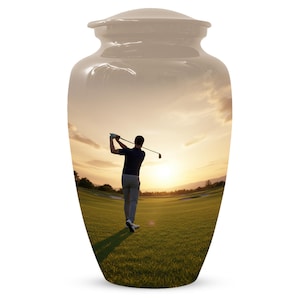Può includere: Urna in ceramica beige con coperchio, con una scena di campo da golf. L'immagine mostra un golfista che fa un swing al tramonto. L'urna è un oggetto commemorativo.