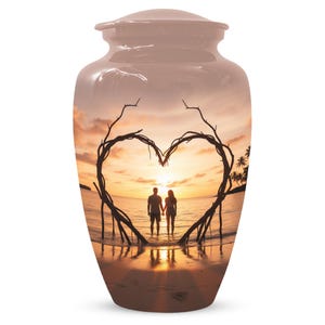 Puede incluir: Urna funeraria de color rosa pálido con una escena de puesta de sol enmarcada por una forma de corazón hecha de ramas de árbol. La imagen muestra a una pareja tomados de la mano en el mar. La urna es cilíndrica con tapa.