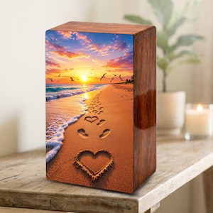 May include: A wooden cremation urn with a glossy finish. The front panel features a beach scene with a sunset, footprints, and a heart drawn in the sand. The sky is filled with birds. The urn is displayed on a wooden surface.