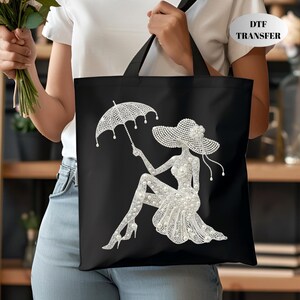 May include: Black tote bag featuring a rhinestone design of a woman in a hat holding an umbrella. The woman is wearing a dress and high heels. The bag has black handles and the text "DTF TRANSFER" in the upper right corner.