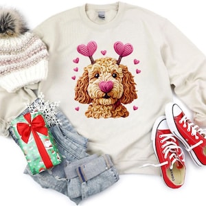 May include: Cream-colored sweatshirt featuring a cartoon dog with heart-shaped antlers and a pink heart nose. Small pink hearts surround the dog. Red Converse sneakers, a white knit hat with a pom-pom, and a wrapped gift with a red bow are also in the image.