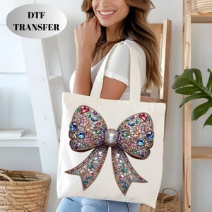 May include: Cream-colored tote bag featuring a large, ornate bow design. The bow is embellished with colorful gemstones in shades of red, blue, pink, and green. The text "DTF TRANSFER" is in a white oval at the top.