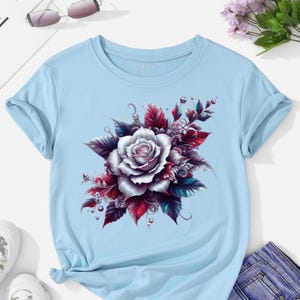 May include: Light blue t-shirt featuring a large, detailed floral design. The graphic showcases a white rose surrounded by burgundy, dark blue, and green leaves. The shirt has short sleeves and a round neckline, with the sleeves rolled up.