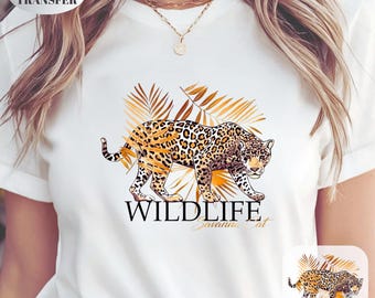 May include: White t-shirt featuring a leopard design with the text "WILDLIFE Savannah Cat". The leopard is depicted with a spotted pattern and is set against a backdrop of golden-brown leaves.