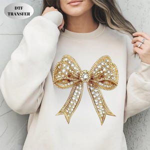 May include: Cream-colored sweatshirt featuring a large, ornate bow design. The bow is gold-colored and encrusted with numerous sparkling rhinestones. The text "DTF TRANSFER" is in a silver oval in the upper left corner.