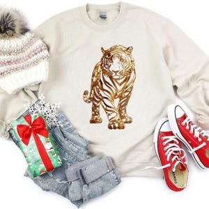 May include: Cream-colored sweatshirt featuring a gold tiger design. The image also includes a white and gray knit hat with a pom-pom, a wrapped gift with a red bow, a pair of blue jeans, and red Converse sneakers.