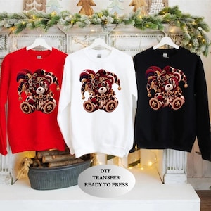 May include: Three crewneck sweatshirts in red, white, and black, each featuring a graphic of a teddy bear wearing a jester hat and collar with gold accents. A white oval sign in the foreground displays the text DTF TRANSFER READY TO PRESS.