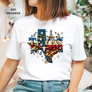 May include: White t-shirt featuring a colorful Texas-shaped graphic. The graphic incorporates the state flag, cowboy hats, a longhorn skull, and an oil derrick. The text "TEXAS" is prominently displayed within the design.