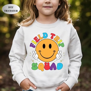 May include: A white sweatshirt featuring a large, colorful graphic. The graphic includes a smiling face with peace signs and the words "FIELD TRIP SQUAD" in rainbow colors. The sweatshirt is a casual style.