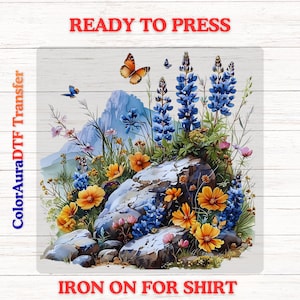 May include: A square DTF transfer sheet featuring a mountain landscape with blue lupine flowers, orange cosmos, and butterflies. The text READY TO PRESS appears at the top, ColorAuraDTF Transfer on the left, and IRON ON FOR SHIRT at the bottom.