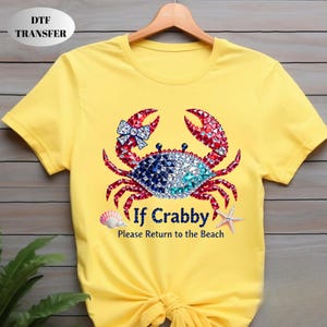 May include: Yellow t-shirt featuring a crab design with red, blue, and white jewel-like accents. The crab has a bow and is surrounded by a seashell and starfish. Text reads "If Crabby Please Return to the Beach."