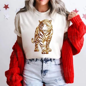 May include: Cream-colored t-shirt featuring a gold tiger design. The tiger is depicted in a detailed, artistic style. The shirt is paired with light-wash denim shorts and a red cardigan.