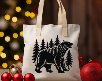 May include: A cream-colored tote bag with a black graphic of a bear and pine trees. The bag has beige handles and is sitting on a wooden surface. Red Christmas ornaments are in the foreground, with a blurred background of Christmas lights.