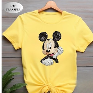 May include: A bright yellow short-sleeved t-shirt featuring a graphic of Mickey Mouse with a sparkling, rhinestone-like texture. The character is depicted with black ears, a tan face, and white gloves, resting a hand on a chin. Text reads DTF TRANSFER.