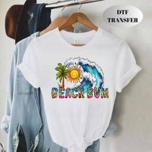 May include: White t-shirt featuring a colorful graphic of a sun, palm tree, and ocean wave. The text "BEACH BUM" is printed in a vibrant, multi-colored font. The shirt is displayed on a hanger.